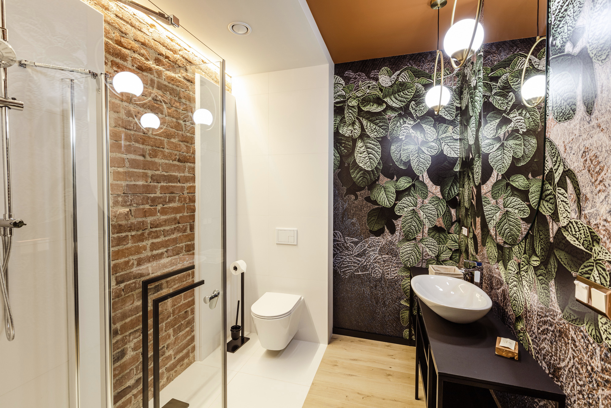 Nowoczesna łazienka z kabiną prysznicową, ceglaną ścianą i tapetą w liście. Umywalka nablatowa na czarnej konsoli. Oświetlenie punktowe i wiszące lampy.