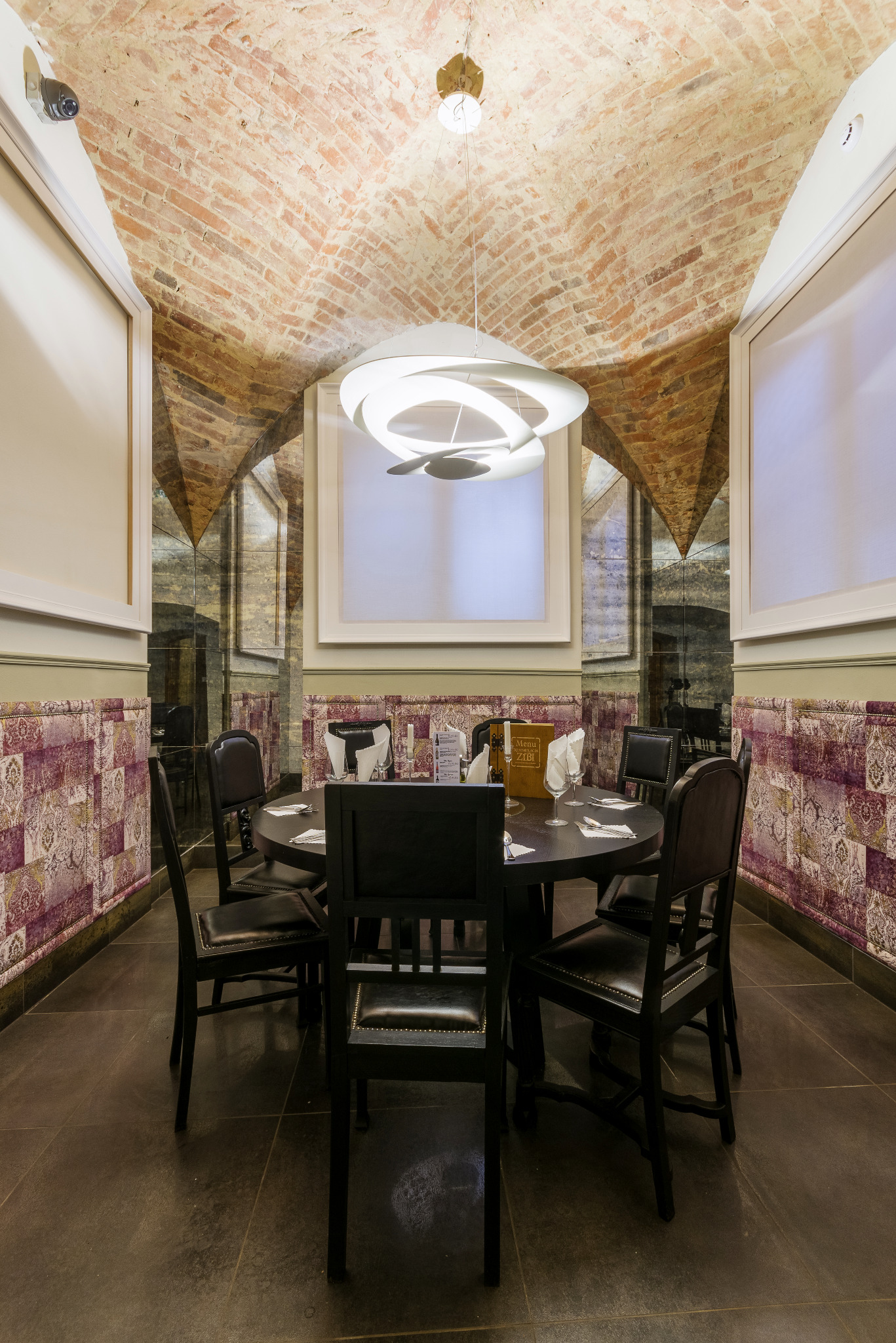 Stylowa aranżacja sali restauracyjnej z okrągłym stołem i krzesłami, ceglanym sklepieniem i nowoczesną lampą. Dekoracyjne panele ścienne i lustra dodają elegancji.