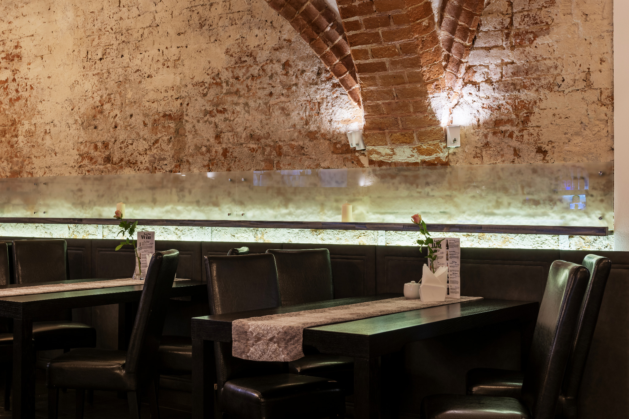 Stylowe wnętrze restauracji z ceglanym łukiem, oświetleniem punktowym i eleganckimi, ciemnymi meblami. Na stołach dekoracje z róż i menu.