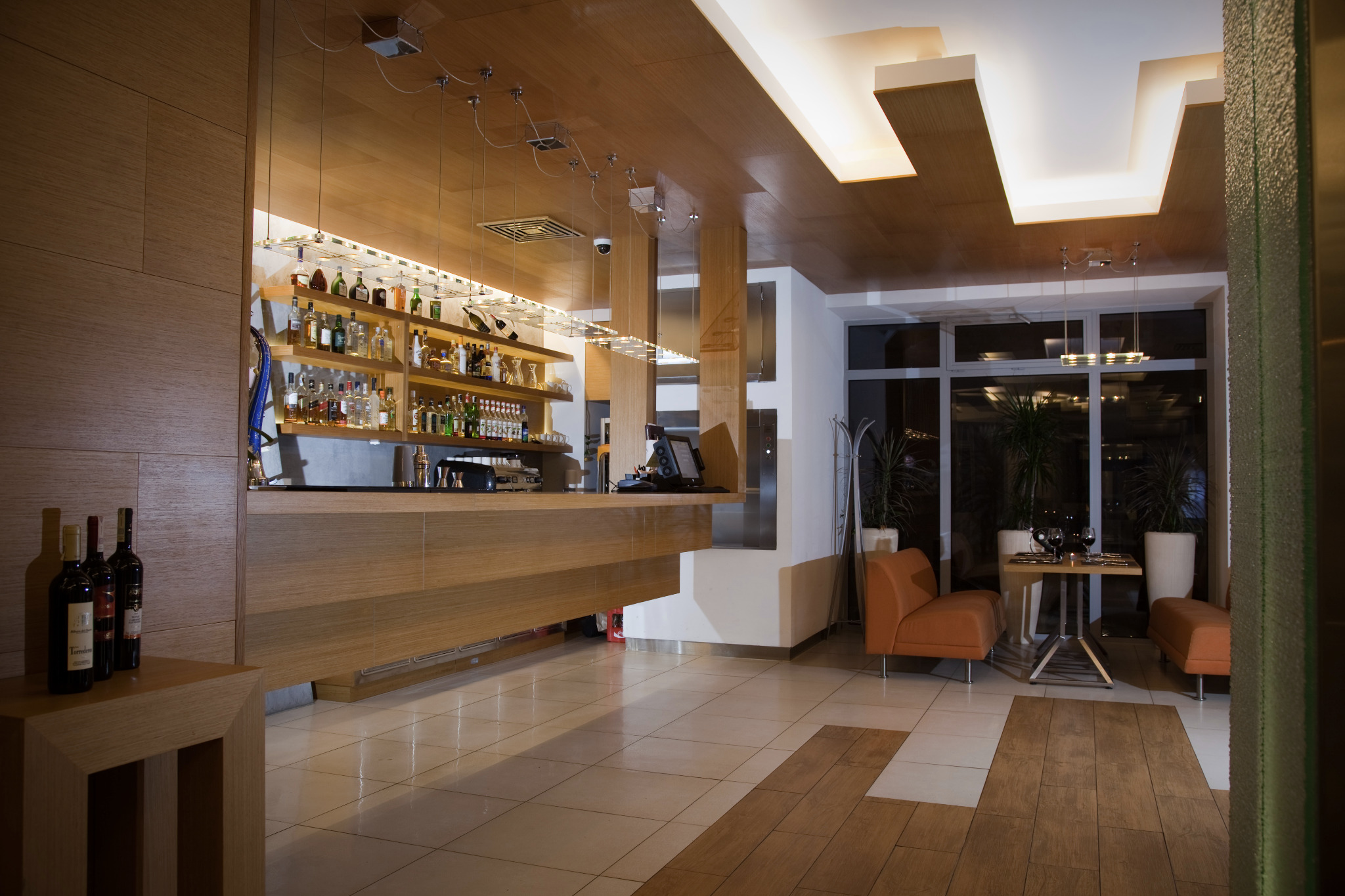 Nowoczesne wnętrze baru z drewnianym wykończeniem, oświetleniem punktowym i butelkami alkoholu na półkach. Widoczne stoliki i krzesła dla gości.