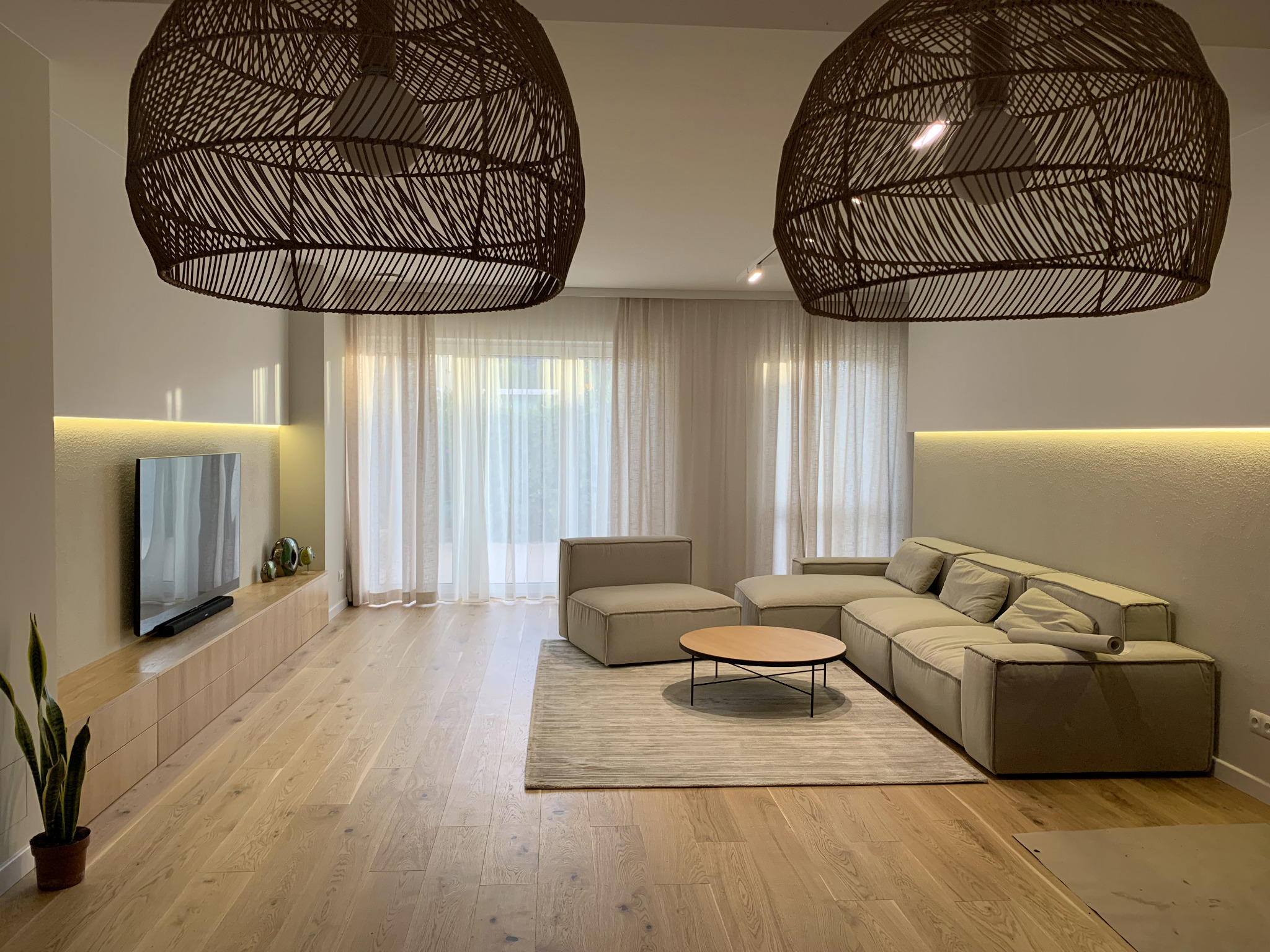 Jasny salon z drewnianą podłogą, beżową sofą modułową, okrągłym stolikiem kawowym i dwoma dużymi, plecionymi lampami wiszącymi. Minimalistyczny design z akcentami naturalnymi.