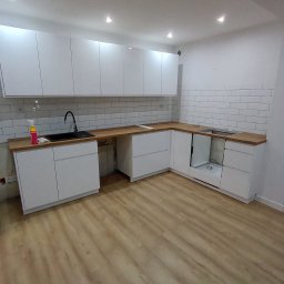 Norbert Gębuś Firma Budowlana LEOŚ - Nowoczesna, biała kuchnia z drewnianym blatem i białymi płytkami na ścianie. Widoczne szafki górne i dolne, zlew z czarną baterią. Podłoga wyłożona panelami w odcieniach beżu.