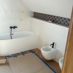 Norbert Gębuś Firma Budowlana LEOŚ - Biała wanna i bidet z czarną armaturą w łazience na poddaszu, zabezpieczone folią i papierem malarskim. Dekoracyjna mozaika nad wanną. Parkiet w jodełkę.