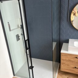 Norbert Gębuś Firma Budowlana LEOŚ - Nowoczesna kabina prysznicowa z czarnymi profilami i deszczownicą, na tle ściany z ciemnych płytek o geometrycznym wzorze. Umywalka z drewnianą szafką i lustro w tle.