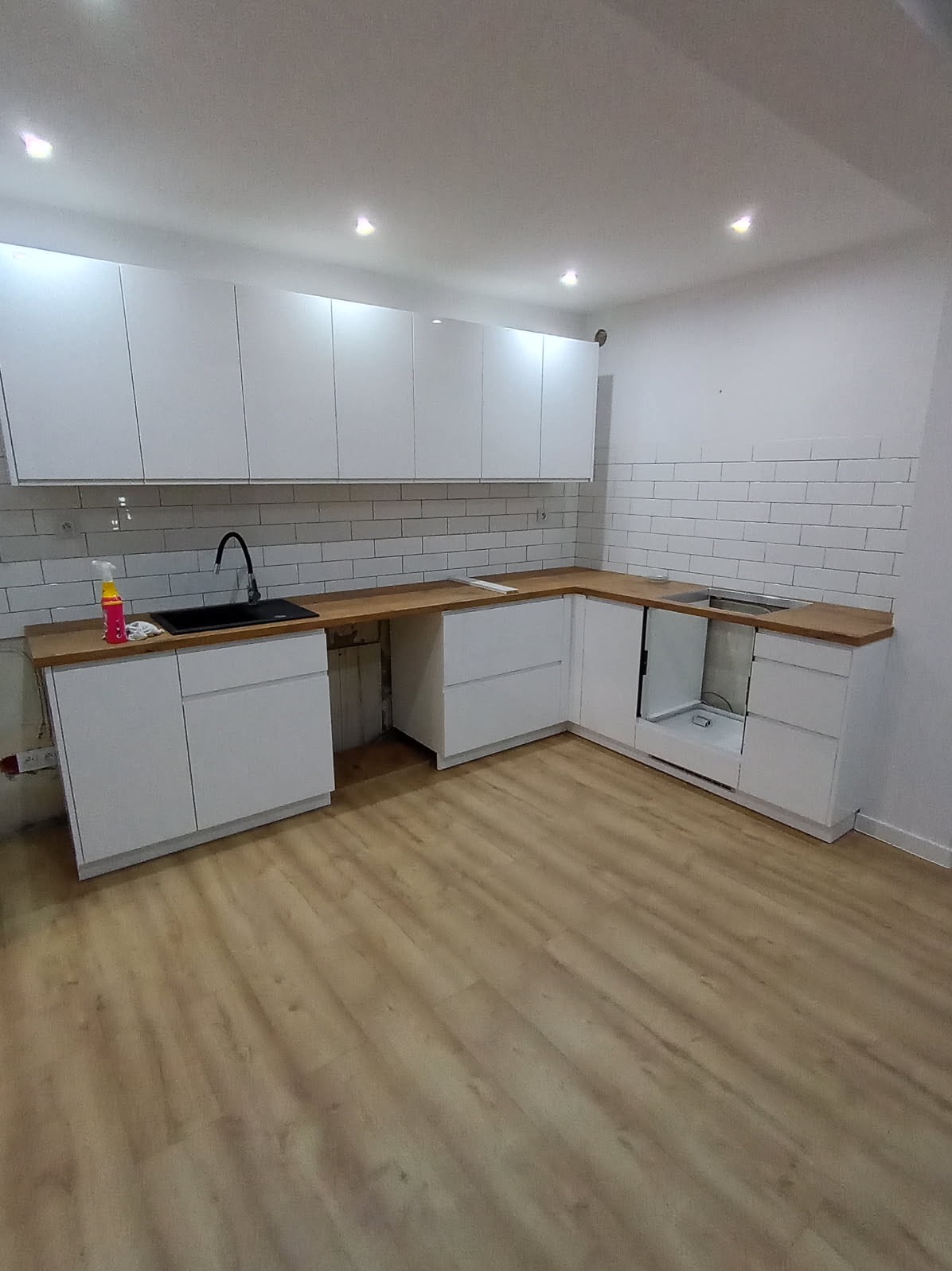 Nowoczesna, biała kuchnia z drewnianym blatem i białymi płytkami na ścianie. Widoczne szafki górne i dolne, zlew z czarną baterią. Podłoga wyłożona panelami w odcieniach beżu.