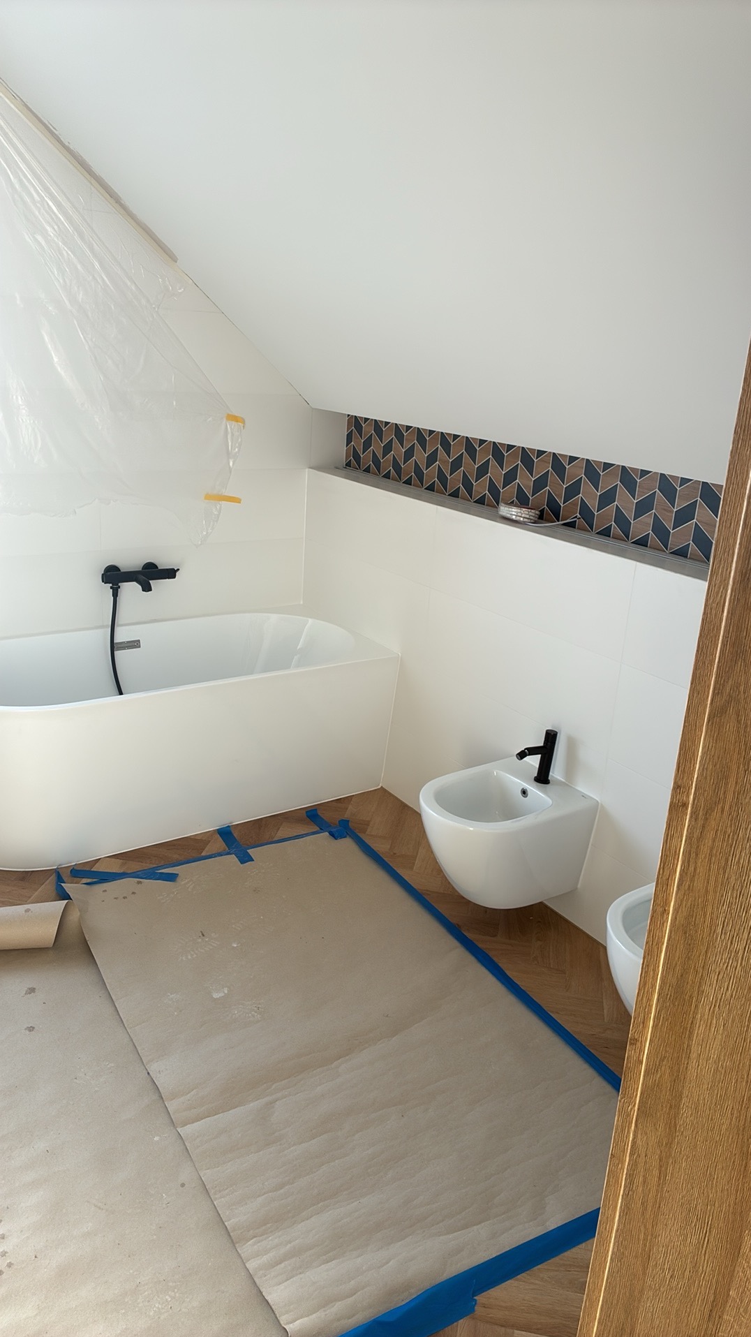 Biała wanna i bidet z czarną armaturą w łazience na poddaszu, zabezpieczone folią i papierem malarskim. Dekoracyjna mozaika nad wanną. Parkiet w jodełkę.