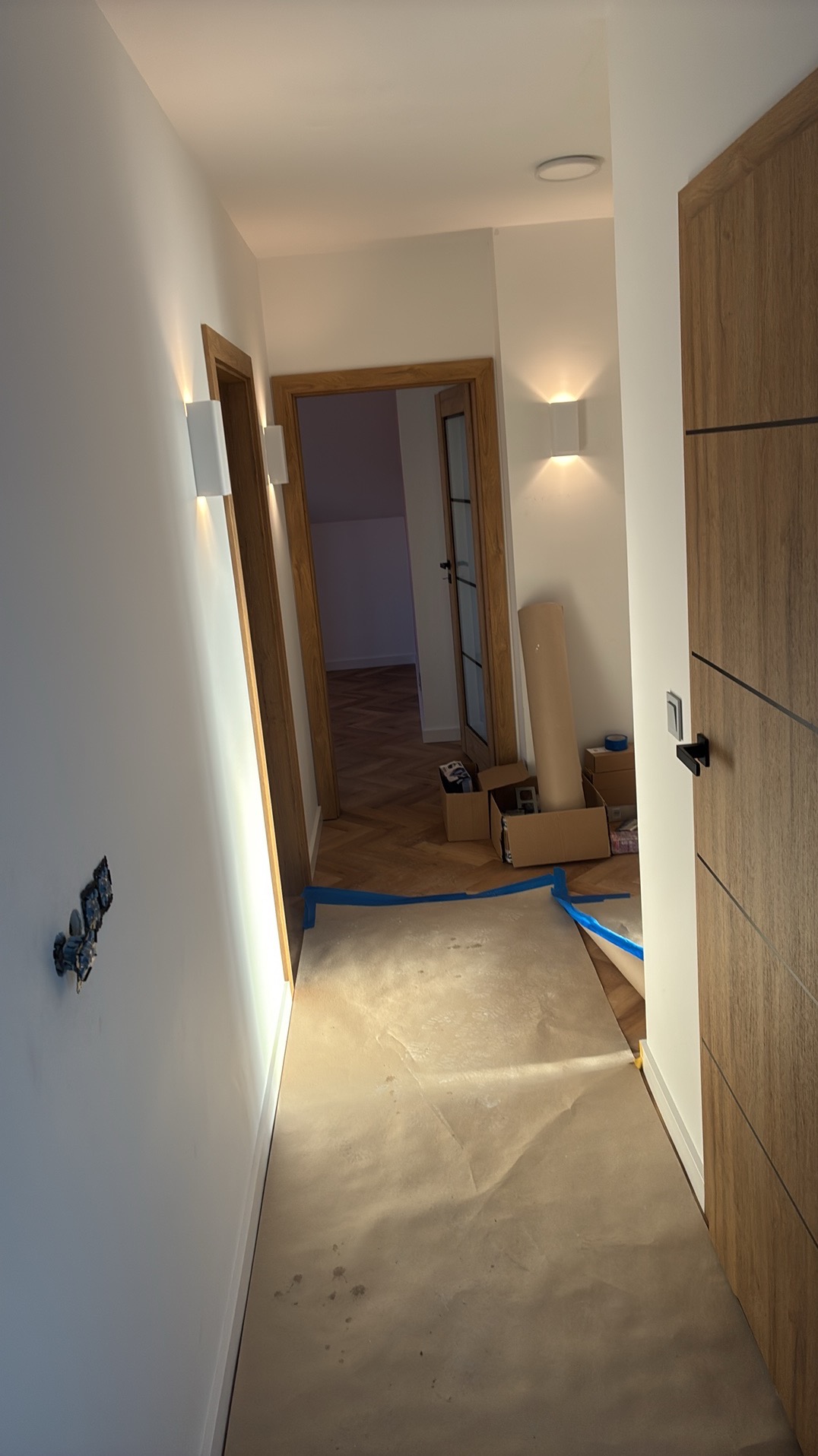 Korytarz w trakcie prac wykończeniowych: zabezpieczona podłoga, widoczne puszki elektryczne i kartony. W tle parkiet i drzwi z ościeżnicami. Nowoczesne oświetlenie.