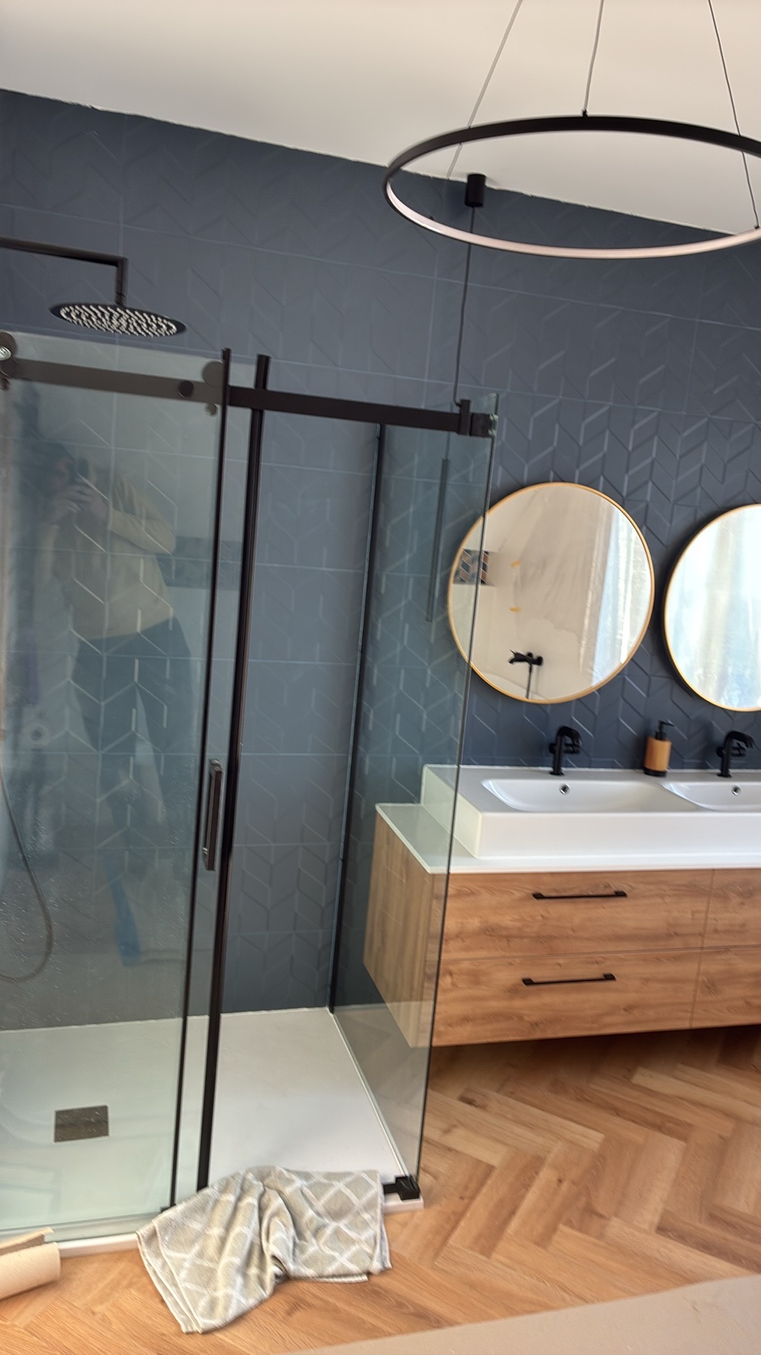 Nowoczesna łazienka z kabiną prysznicową typu walk-in, drewnianą szafką z umywalkami i okrągłymi lustrami na tle ściany z geometrycznym wzorem.
