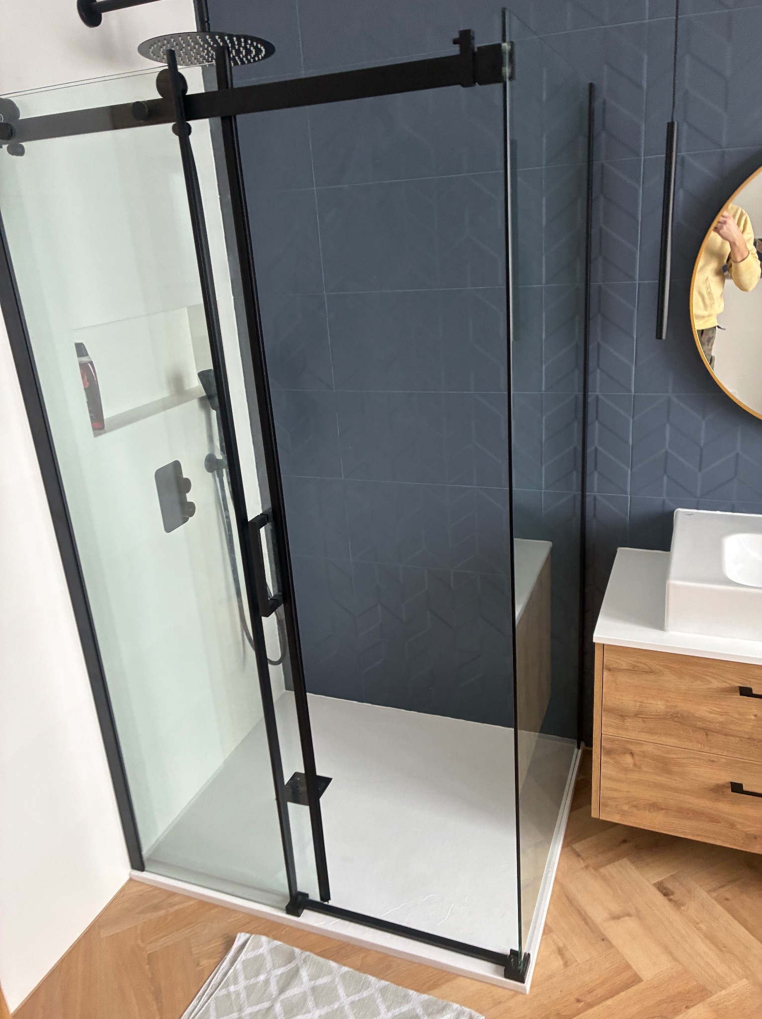 Nowoczesna kabina prysznicowa z czarnymi profilami i deszczownicą, na tle ściany z ciemnych płytek o geometrycznym wzorze. Umywalka z drewnianą szafką i lustro w tle.