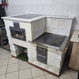 ROMAN WALKOWSKI - Biały, kaflowy piec kuchenny z żeliwną płytą grzewczą i piekarnikiem, widoczny w pomieszczeniu z wykafelkowanymi ścianami i podłogą. Detal wykończenia w stylu retro.
