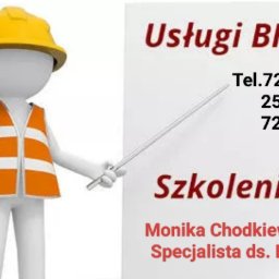 Monika Chodkiewicz - Manekin w kasku i kamizelce BHP wskazuje na tablicę z napisem 'Usługi BHP Szkolenia' i danymi kontaktowymi Moniki Chodkiewicz, specjalisty ds. BHP.