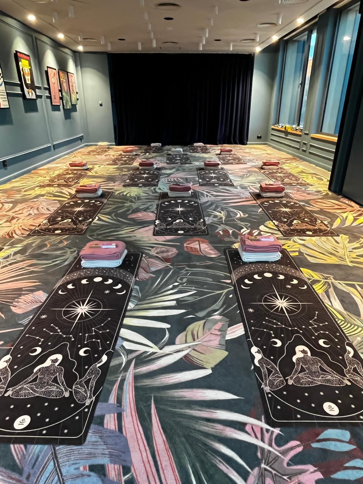 Sala do jogi z rozłożonymi matami z motywem astrologicznym i ręcznikami. W tle zasłony i okna. Podłoga pokryta wzorzystym dywanem w tropikalne liście.