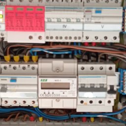 Usługi Elektryczne - Rozdzielnia elektryczna z zabezpieczeniami, licznikiem i przewodami w kolorach: niebieskim, żółto-zielonym, brązowym. Widoczne bezpieczniki i listwy zaciskowe.