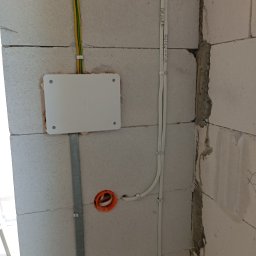 Usługi Elektryczne - Biała puszka elektryczna z przewodami w ścianie z betonu komórkowego. Widoczne kable prowadzone po ścianie i otwór pod gniazdko. Etap prac instalacyjnych.