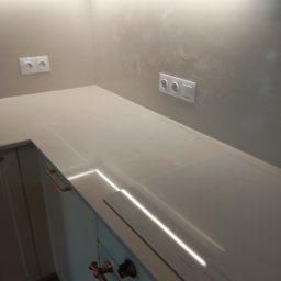 Usługi Elektryczne - Nowoczesna kuchnia z oświetleniem LED podszafkowym i gniazdkami elektrycznymi. Jasny blat i fronty szafek z eleganckimi, złotymi uchwytami.