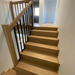 Europejska Grupa Schodów - Jasne, drewniane schody z czarną balustradą w nowym domu. Widok z dołu, w tle widoczne pomieszczenia w stanie surowym.