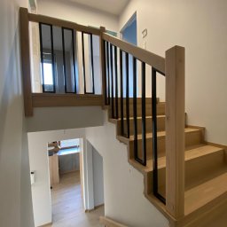 Europejska Grupa Schodów - Jasne, drewniane schody z czarną, metalową balustradą w nowoczesnym wnętrzu. Widok z góry, na dole widoczny fragment podłogi i pomieszczenia.