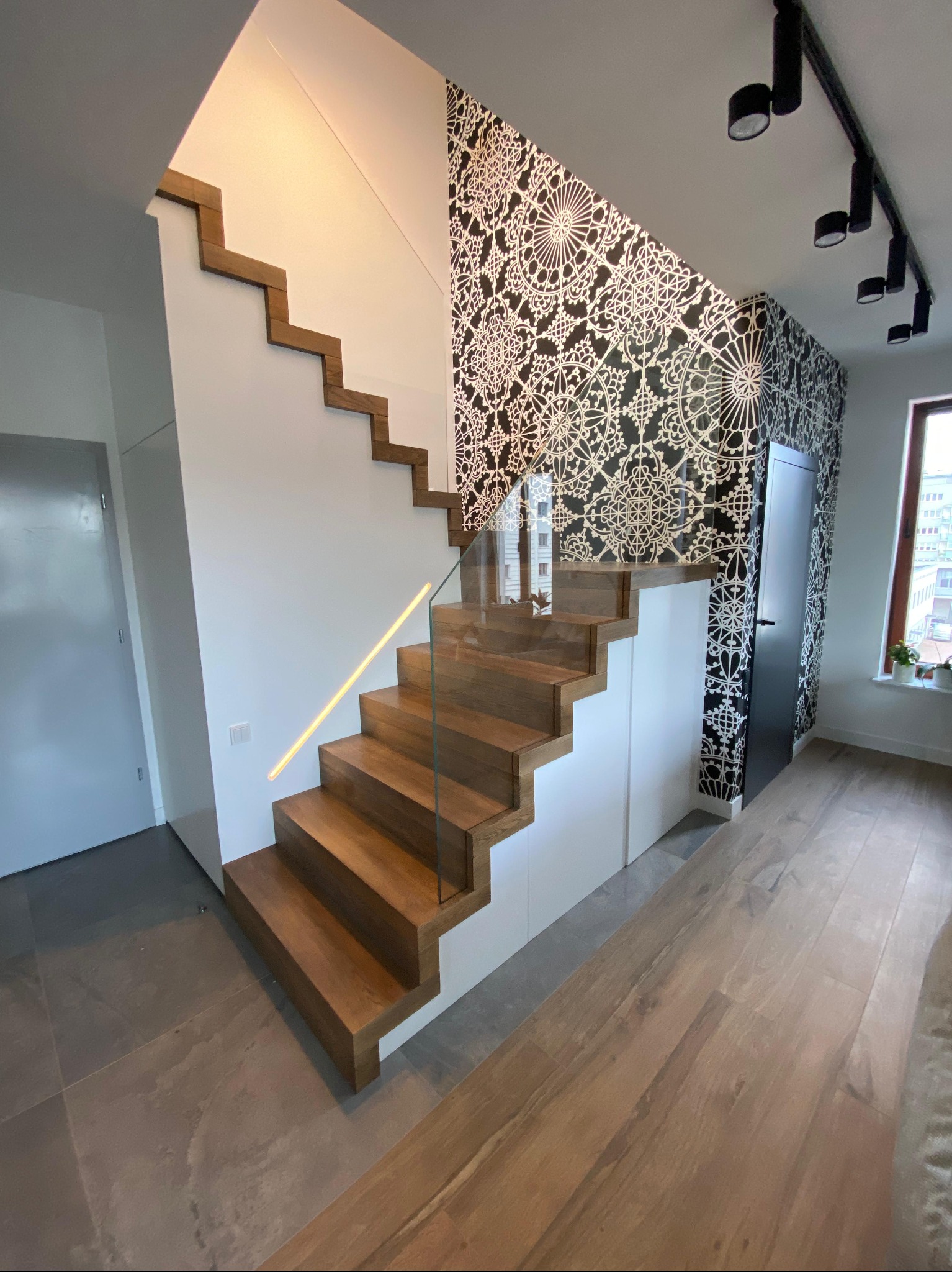 Nowoczesne schody z drewna z podświetleniem LED i szklaną balustradą, na tle ściany z wzorzystą tapetą. Minimalistyczny design, jasne wnętrze.