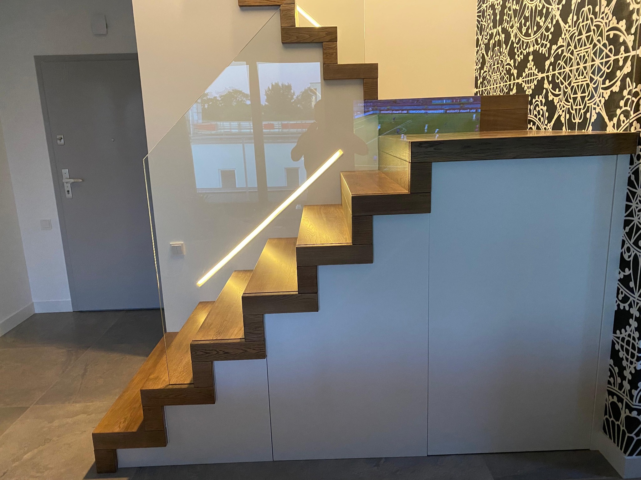 Nowoczesne schody z drewnianymi stopniami, szklaną balustradą i subtelnym oświetleniem LED, wkomponowane w zabudowę z białymi frontami i wzorzystą tapetą.