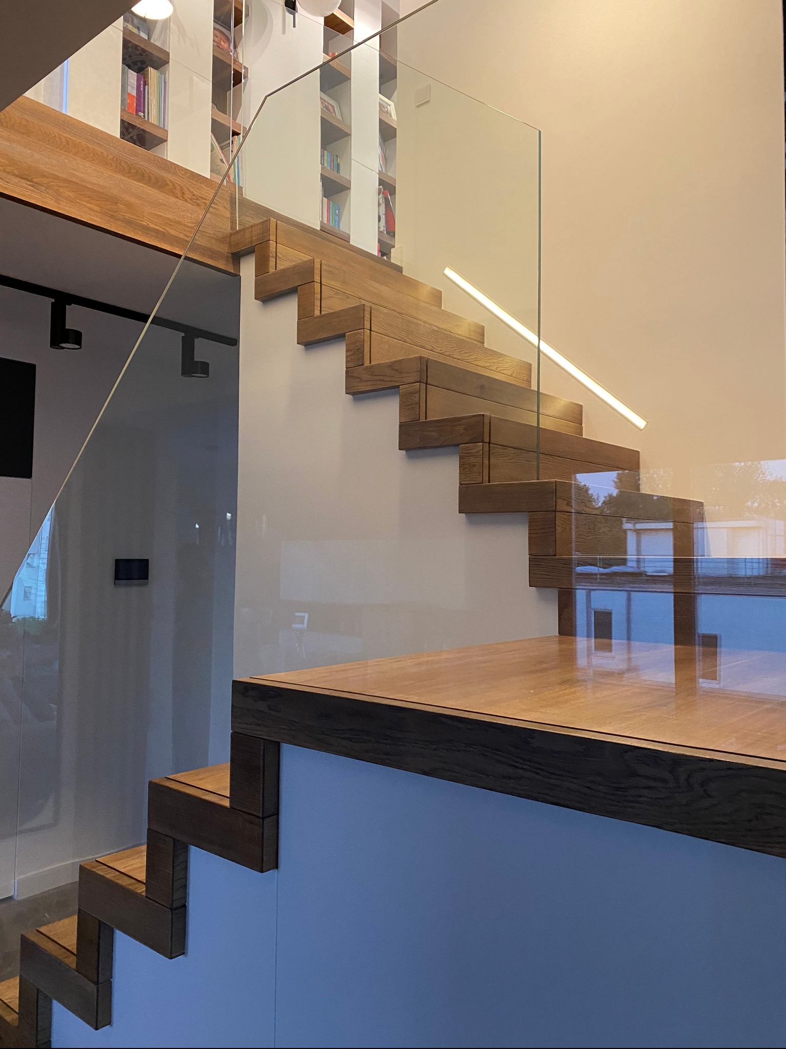 Nowoczesne schody z drewna i szkła z podświetleniem LED, minimalistyczny design, widok z dołu, jasne wnętrze z regałami na książki w tle.