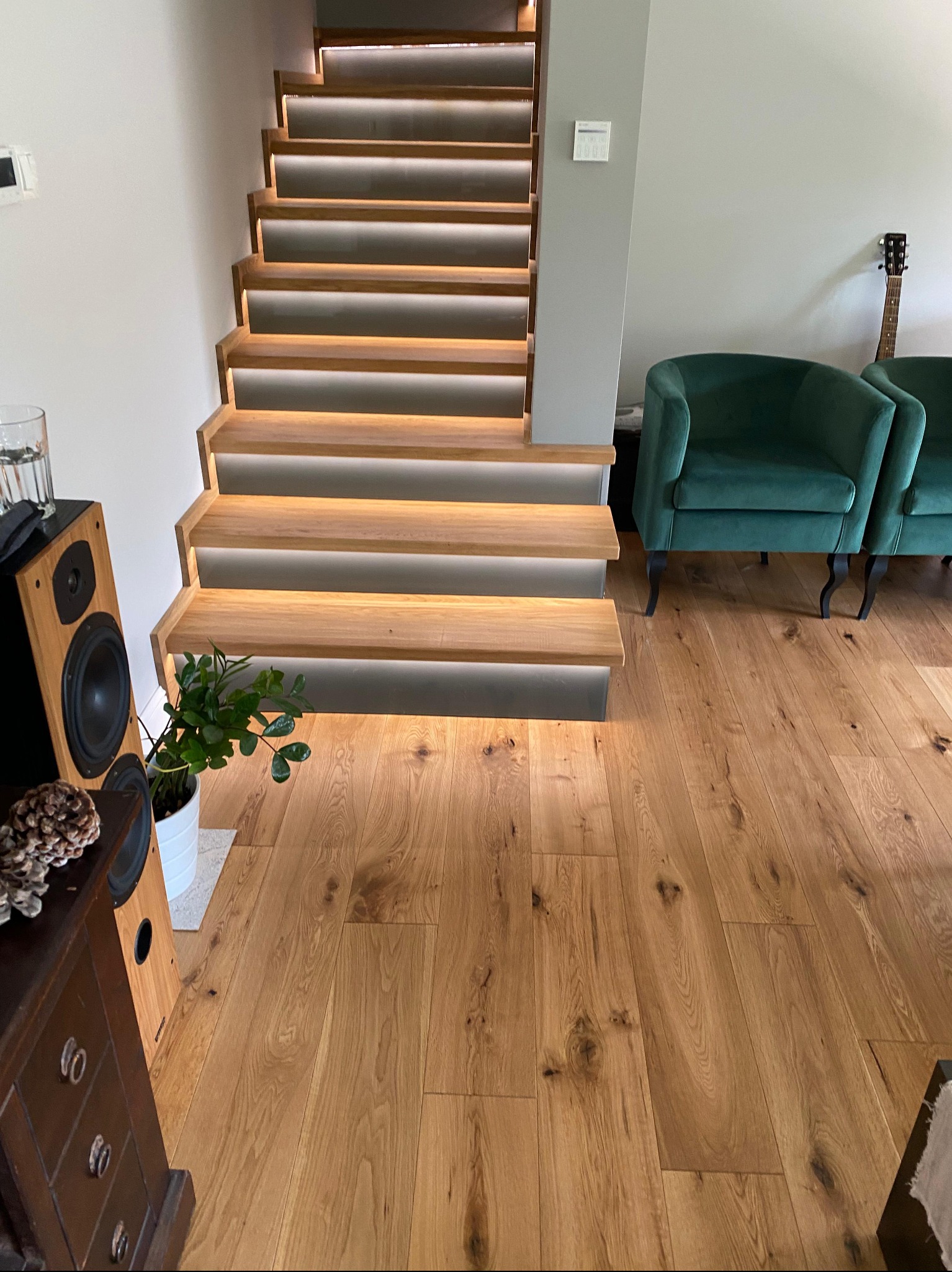 Nowoczesne, podświetlane schody z drewna, zintegrowane z oświetleniem LED, w jasnym wnętrzu z drewnianą podłogą i zielonymi fotelami.