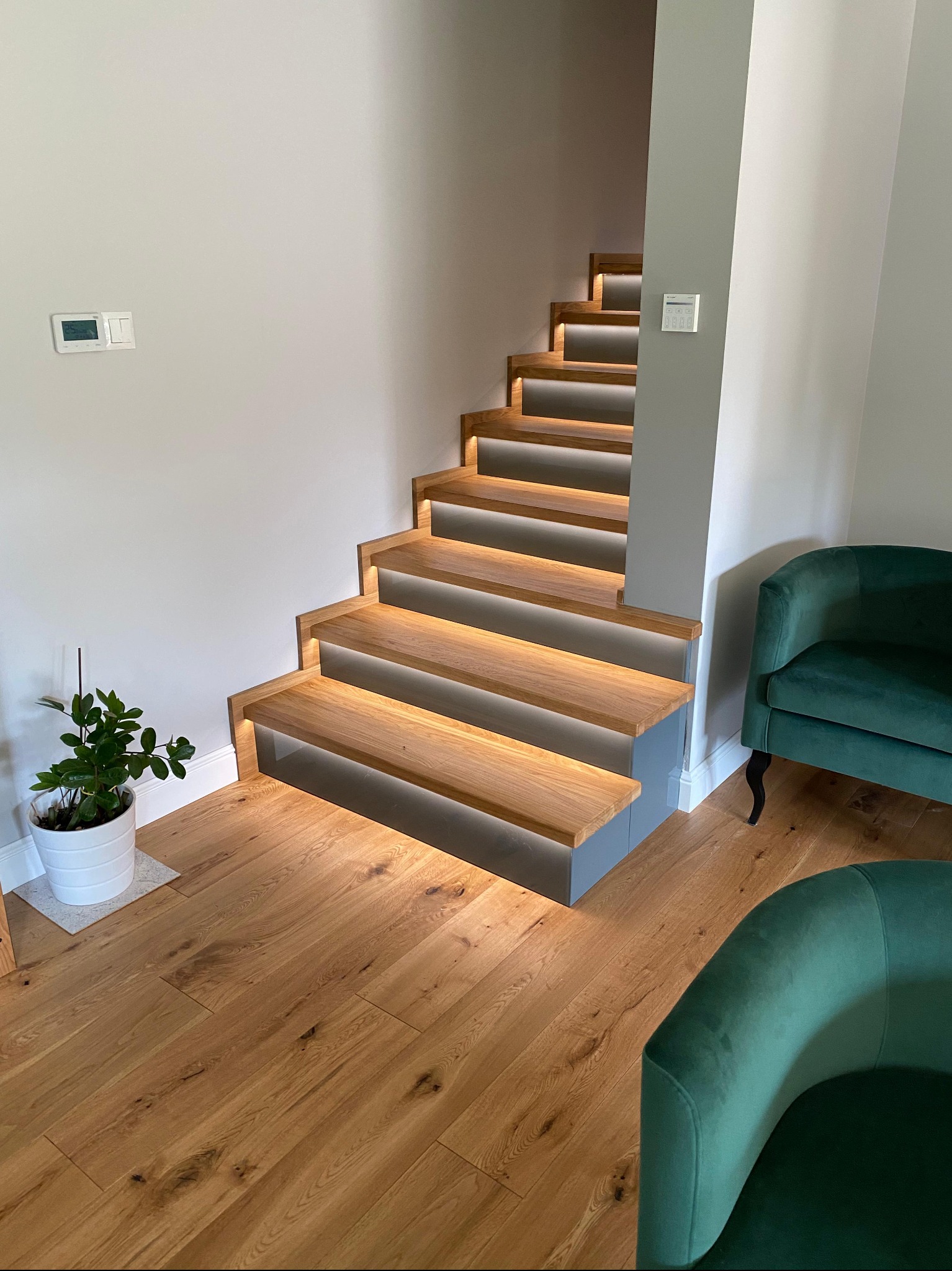 Nowoczesne schody z drewna z subtelnym podświetleniem LED, prowadzące do góry. Obok zielony fotel i roślina doniczkowa. Minimalistyczny design.