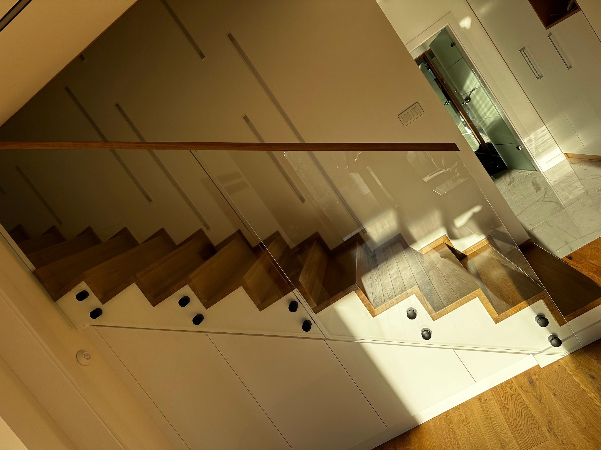 Nowoczesne schody z drewnianymi stopniami i szklaną balustradą, mocowane na białej konstrukcji z szafkami. Widok z góry, ciepłe oświetlenie.