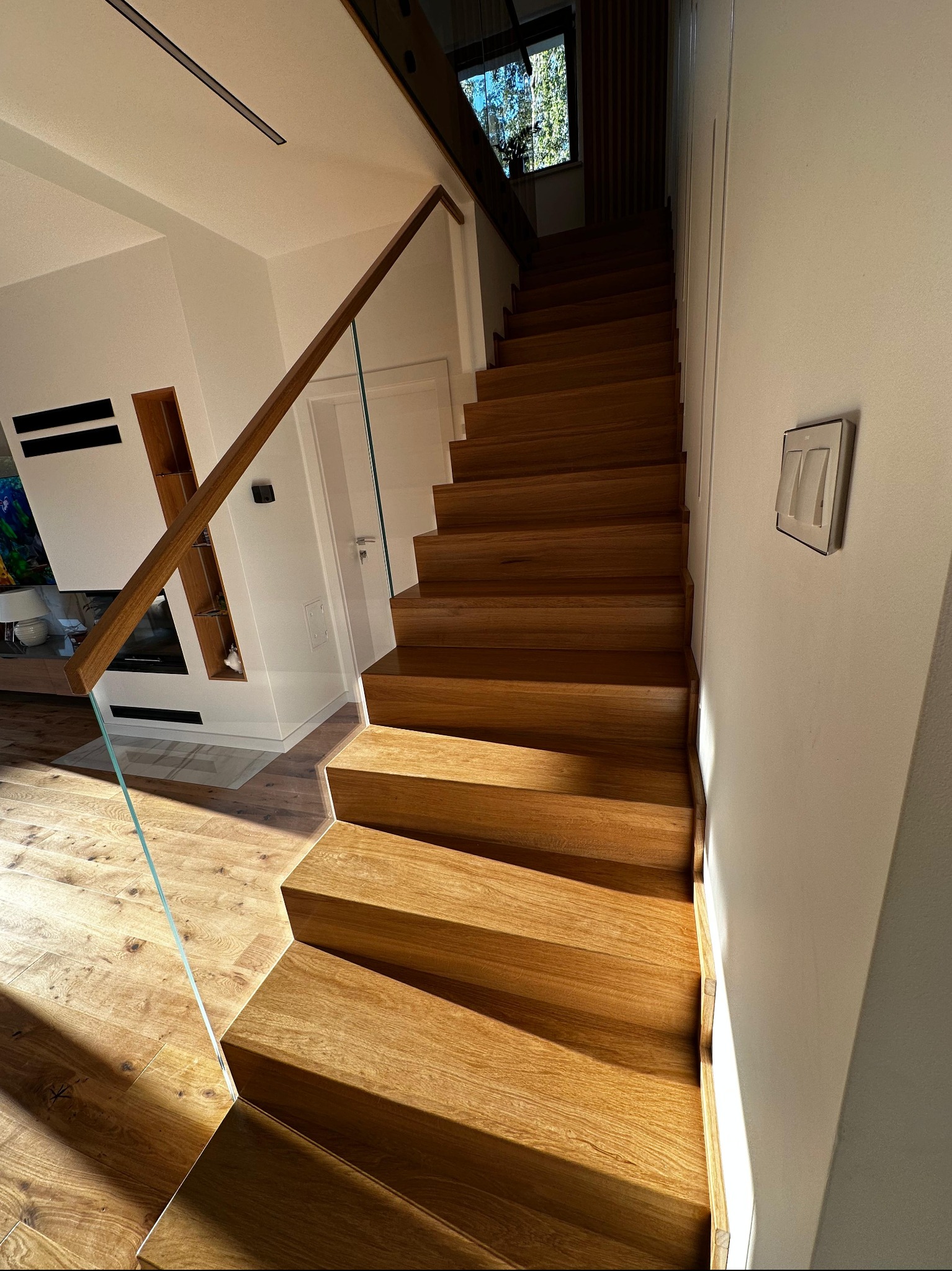 Nowoczesne schody z drewnianymi stopniami i szklaną balustradą, oświetlone naturalnym światłem, harmonijnie komponujące się z wnętrzem domu.