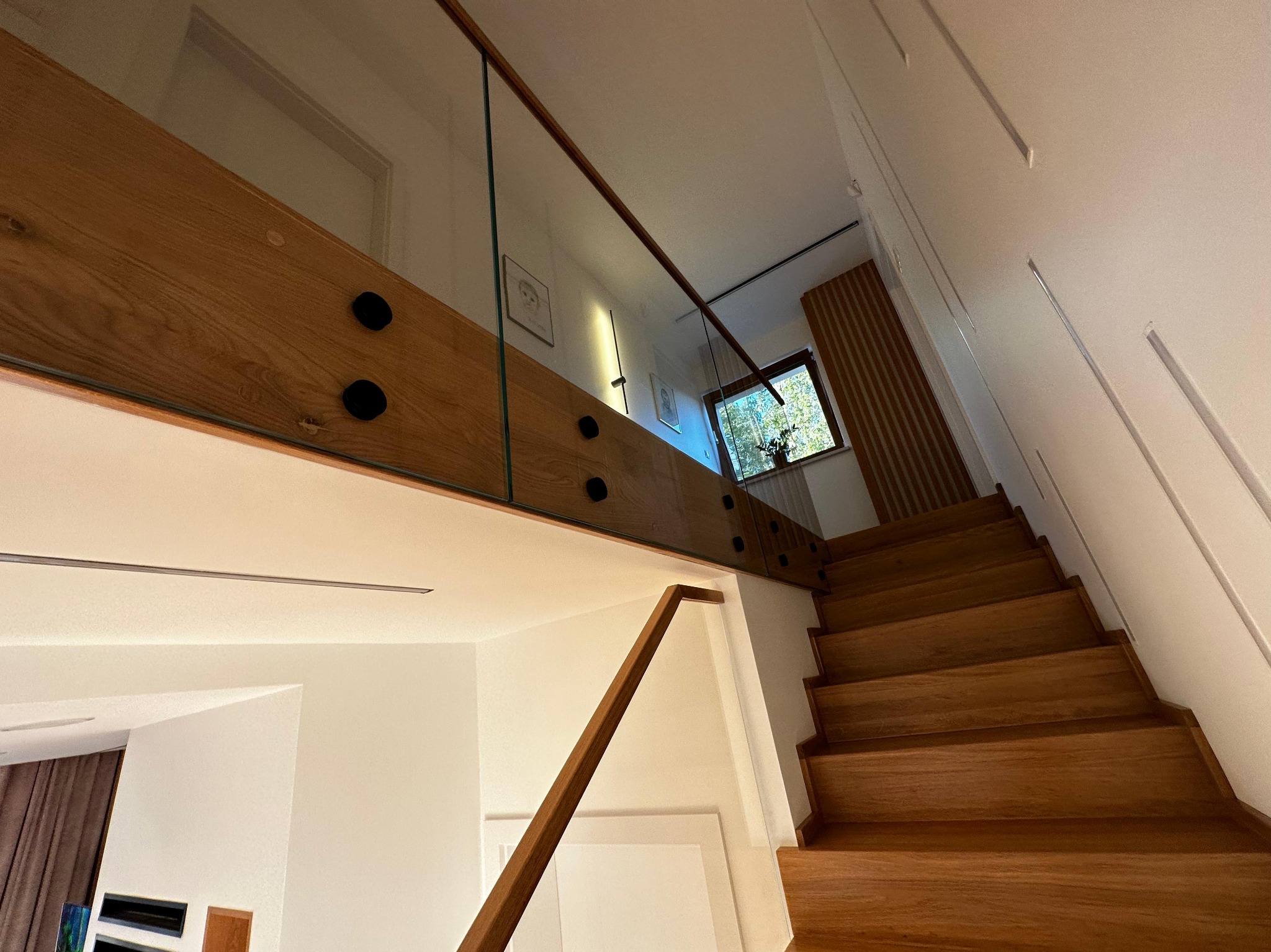 Nowoczesne schody z drewna z balustradą ze szkła, widok z dołu, jasne wnętrze z oknem w tle. Minimalistyczny design, ciepłe oświetlenie.