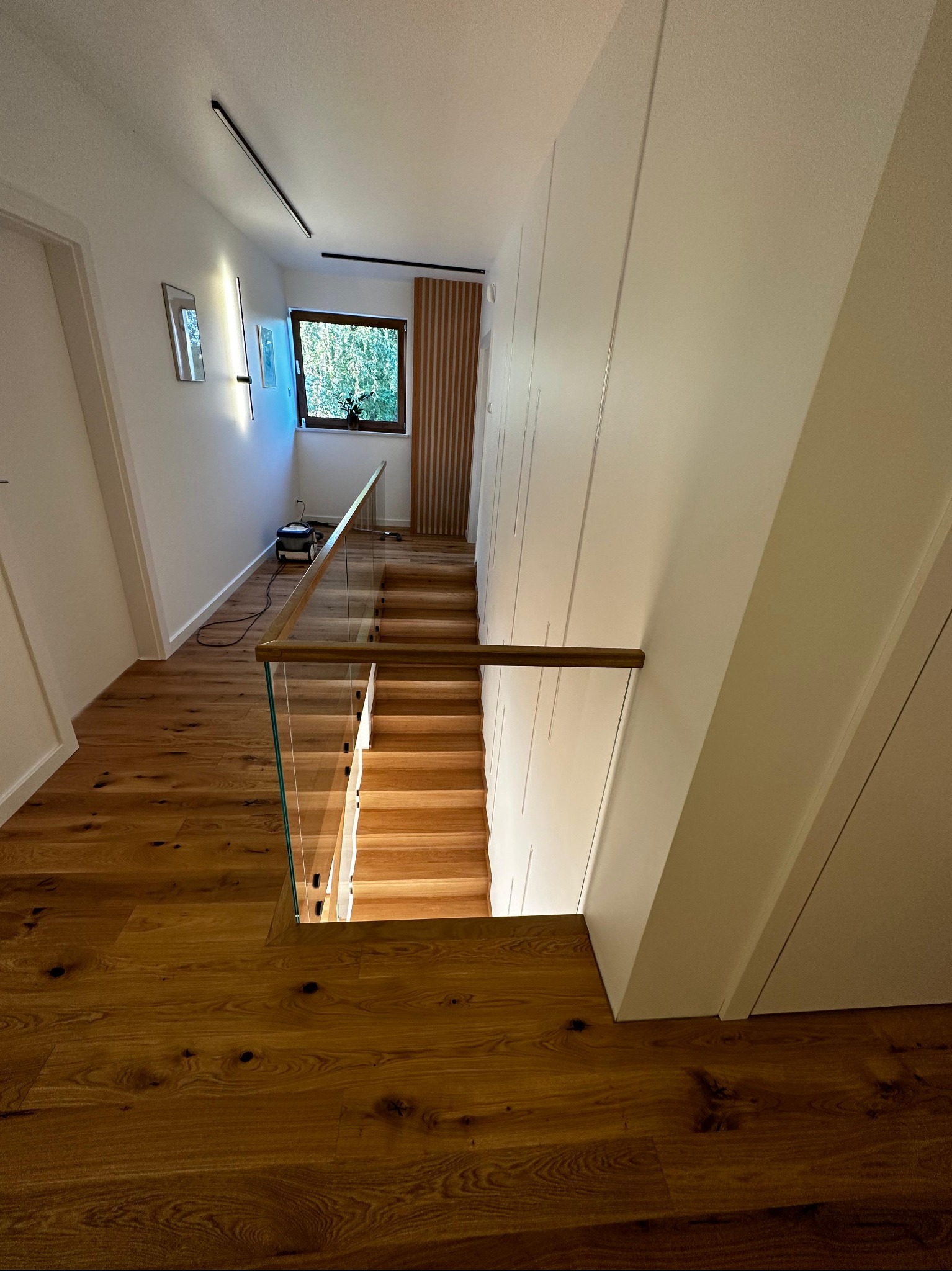 Nowoczesne schody z drewnianymi stopniami i szklaną balustradą, widok z góry, ciepłe oświetlenie, parkiet z wyraźnym usłojeniem, minimalistyczny design.