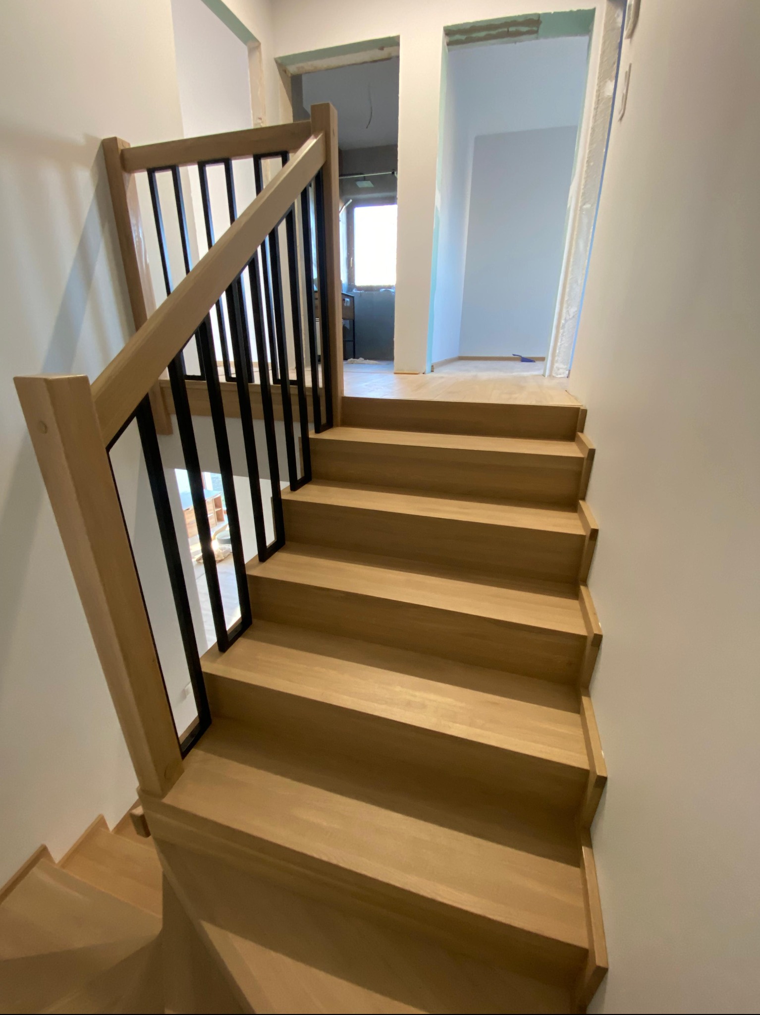 Jasne, drewniane schody z czarną balustradą w nowym domu. Widok z dołu, w tle widoczne pomieszczenia w stanie surowym.