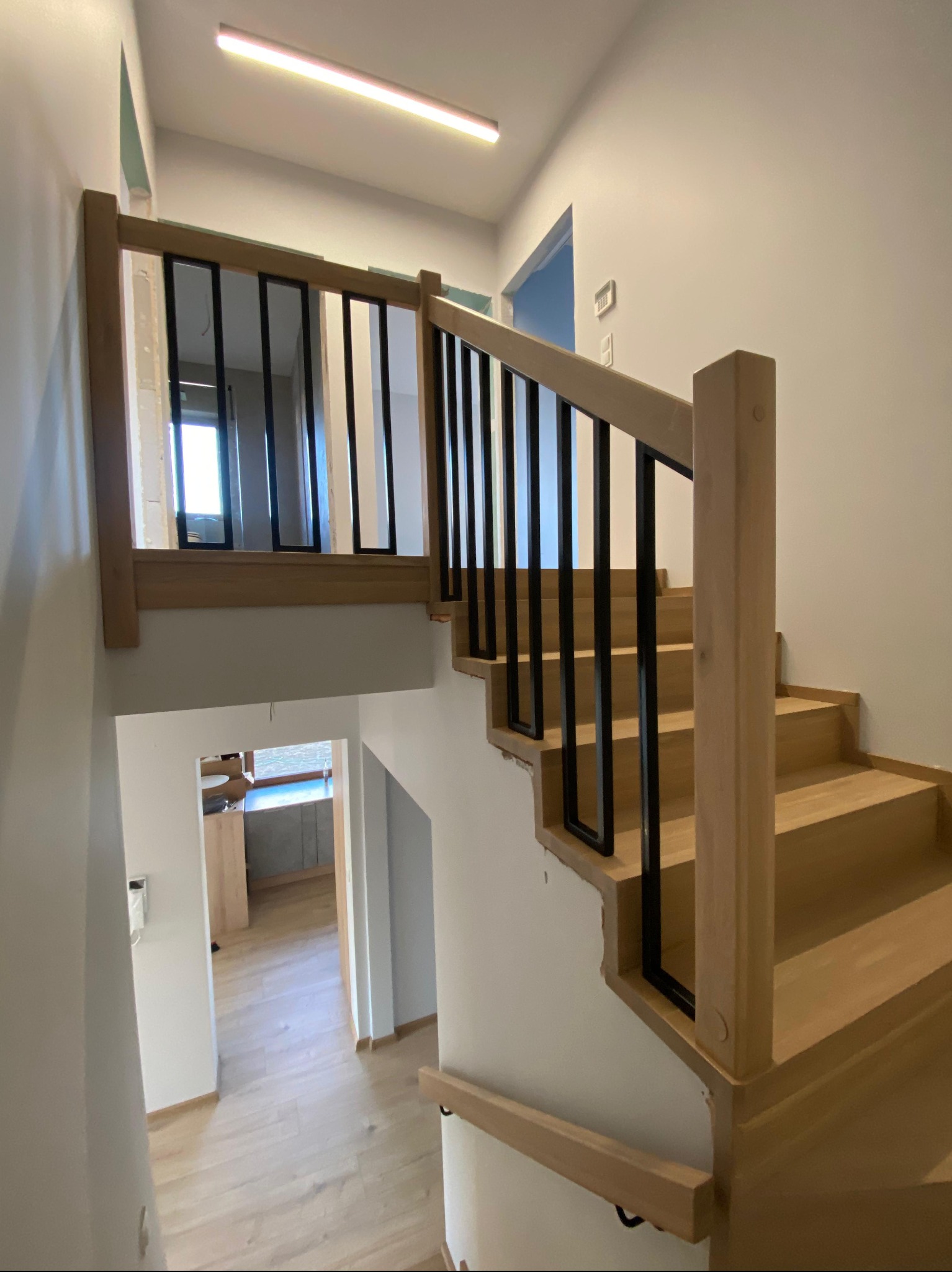 Jasne, drewniane schody z czarną, metalową balustradą w nowoczesnym wnętrzu. Widok z góry, na dole widoczny fragment podłogi i pomieszczenia.