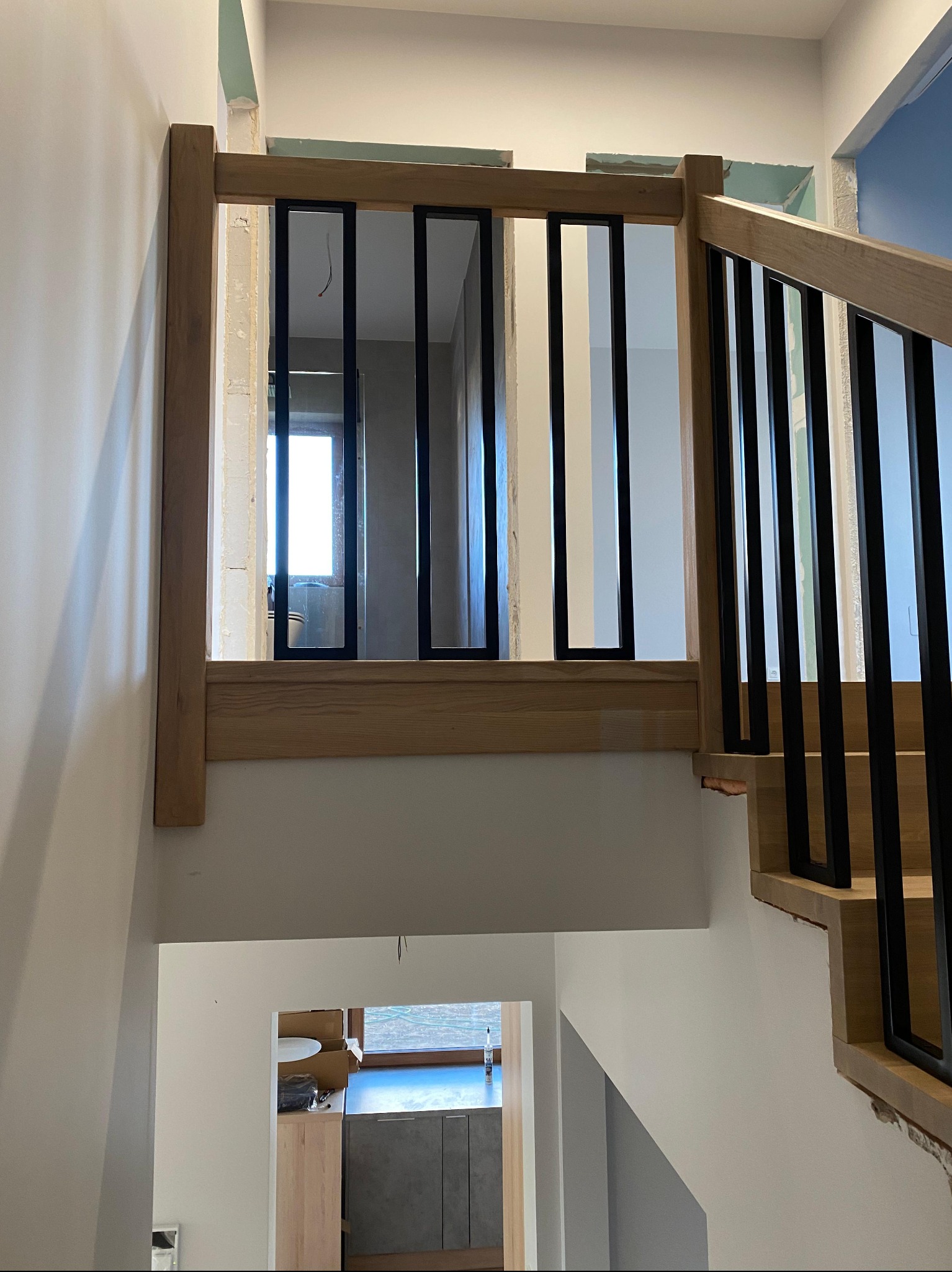 Fragment schodów z drewnianą balustradą i czarnymi, pionowymi elementami. Widok z góry na dół, w tle widoczne pomieszczenia w trakcie wykańczania.