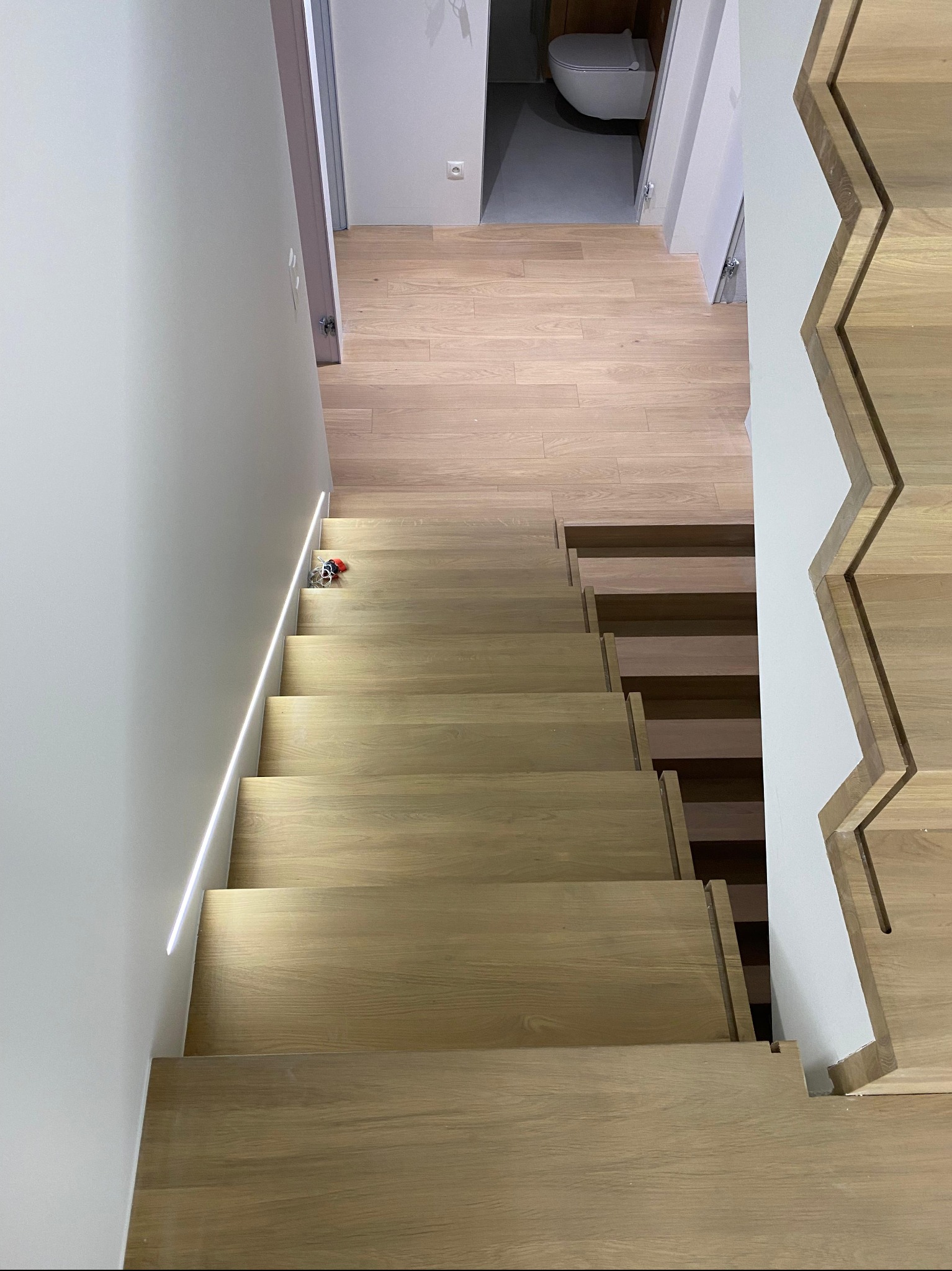 Jasne, drewniane schody z podświetleniem LED wzdłuż ściany. Widok z góry, ukazujący nowoczesny design i precyzję wykonania. W tle widoczna podłoga i fragment łazienki.