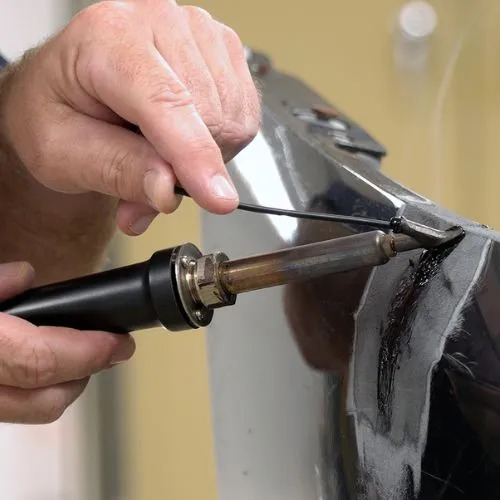 Proces naprawy elementu karoserii z użyciem lutownicy i czarnego tworzywa. Detaliczne ujęcie dłoni i narzędzia w trakcie pracy.