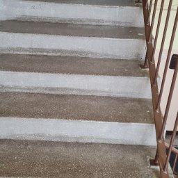 CLEAN PERFECT - Szare, betonowe schody w klatce schodowej z brązową barierką. Widoczne ślady farby na stopniach. Ujęcie z dołu, perspektywa w górę.