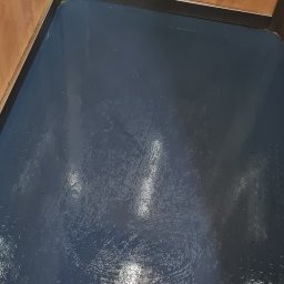 CLEAN PERFECT - Błyszcząca, ciemnoniebieska podłoga po umyciu w rogu pomieszczenia z drewnianymi ścianami i metalowymi listwami przypodłogowymi. Widoczne ślady po mopie.