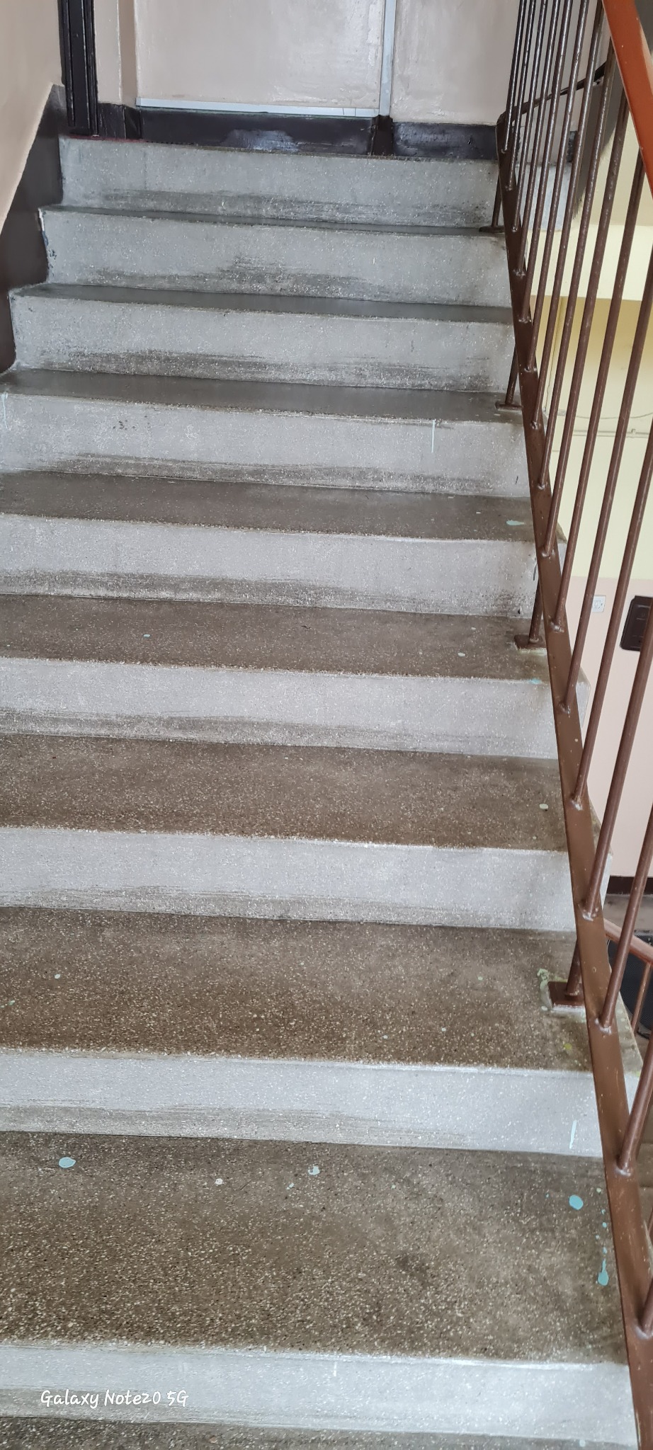 Szare, betonowe schody w klatce schodowej z brązową barierką. Widoczne ślady farby na stopniach. Ujęcie z dołu, perspektywa w górę.