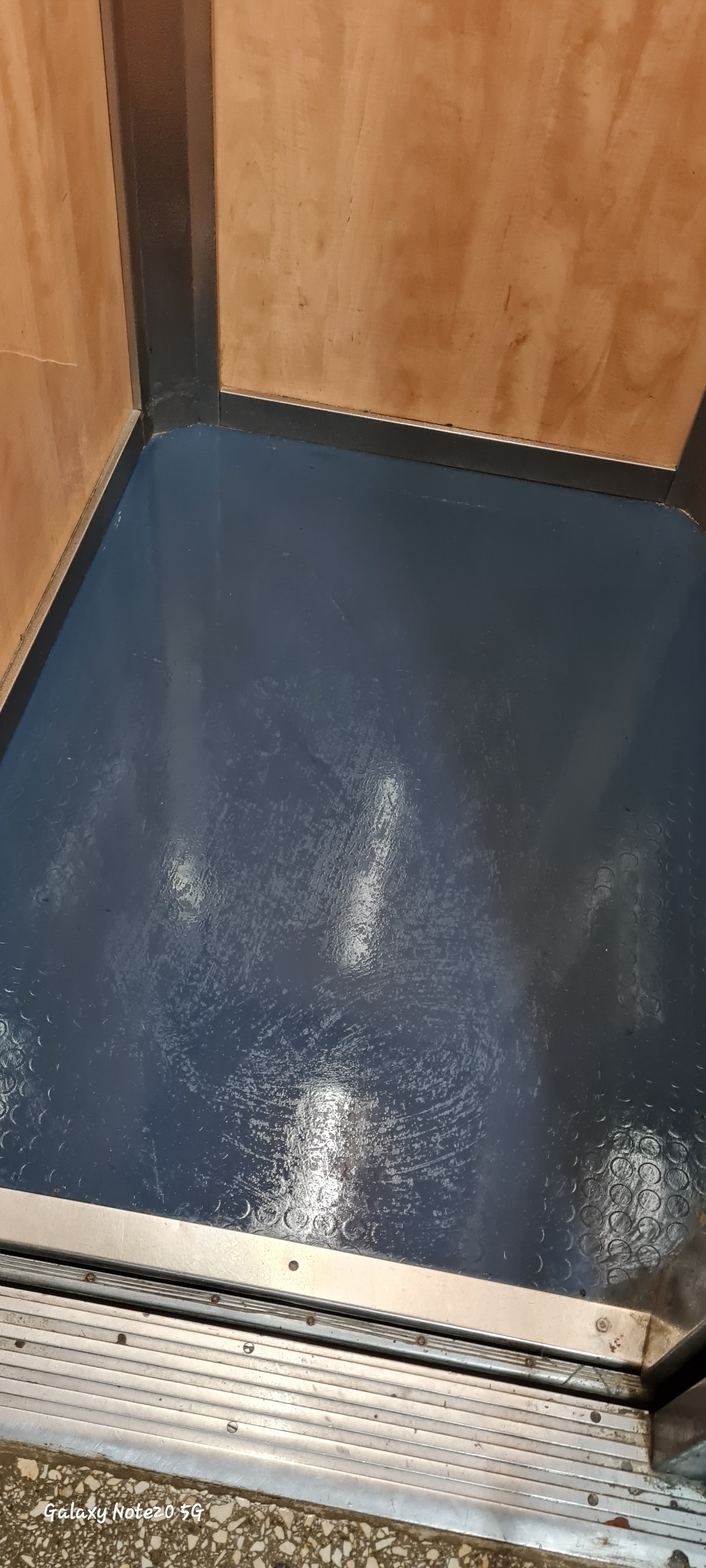 Błyszcząca, ciemnoniebieska podłoga po umyciu w rogu pomieszczenia z drewnianymi ścianami i metalowymi listwami przypodłogowymi. Widoczne ślady po mopie.