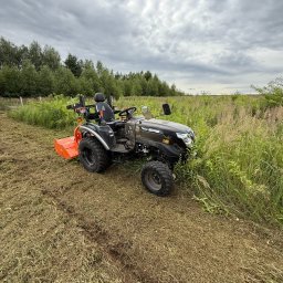 Iskra System Bartłomiej Kiedrzynek - Czarny traktorek Solis z pomarańczowym kosiarką rotacyjną na polu z częściowo skoszoną trawą, w tle drzewa i pochmurne niebo. Widok z boku, lekko z góry.