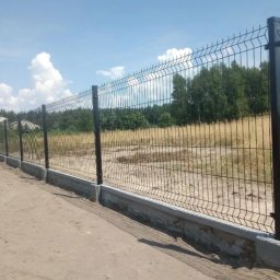 Iskra System Bartłomiej Kiedrzynek - Długi, panelowy płot z siatki z podmurówką betonową, biegnący wzdłuż polnej drogi, z widokiem na zarośnięte pole i drzewa w tle, w słoneczny dzień.