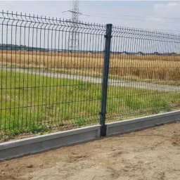 Iskra System Bartłomiej Kiedrzynek - Nowoczesne ogrodzenie panelowe z podmurówką betonową na granicy działki z widokiem na łąkę i słup energetyczny w tle. Estetyczne i funkcjonalne rozwiązanie.