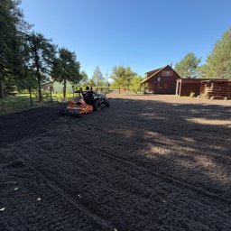 Iskra System Bartłomiej Kiedrzynek - Niwelacja terenu pod trawnik: glebogryzarka wyrównuje ziemię na działce z domem i drzewami w tle, przygotowując podłoże pod założenie ogrodu.
