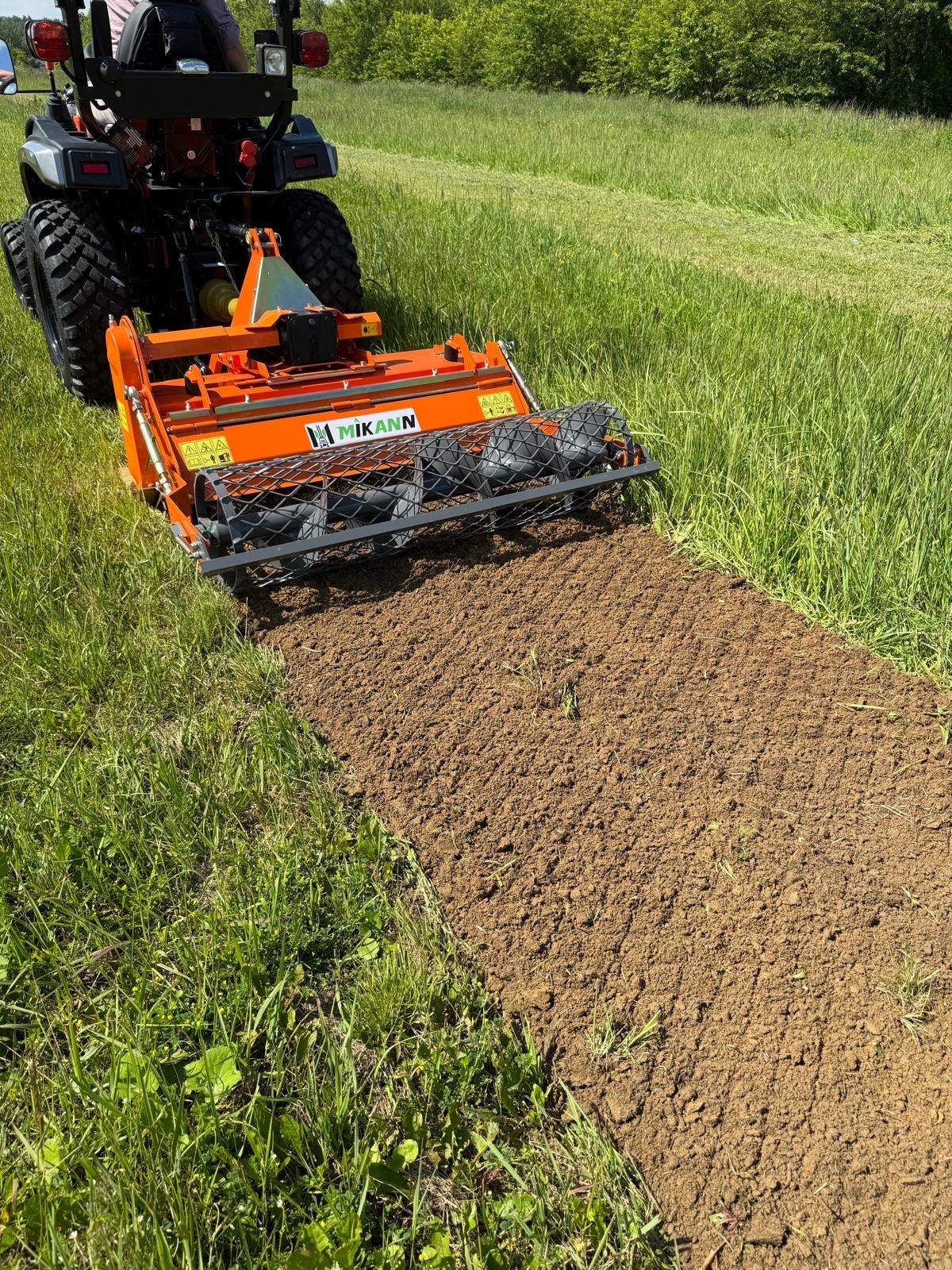 Pomarańczowy traktor z glebogryzarką MIKANN spulchnia ziemię na polu, przygotowując ją pod uprawę. Widok z tyłu maszyny, świeżo obrobiona gleba.