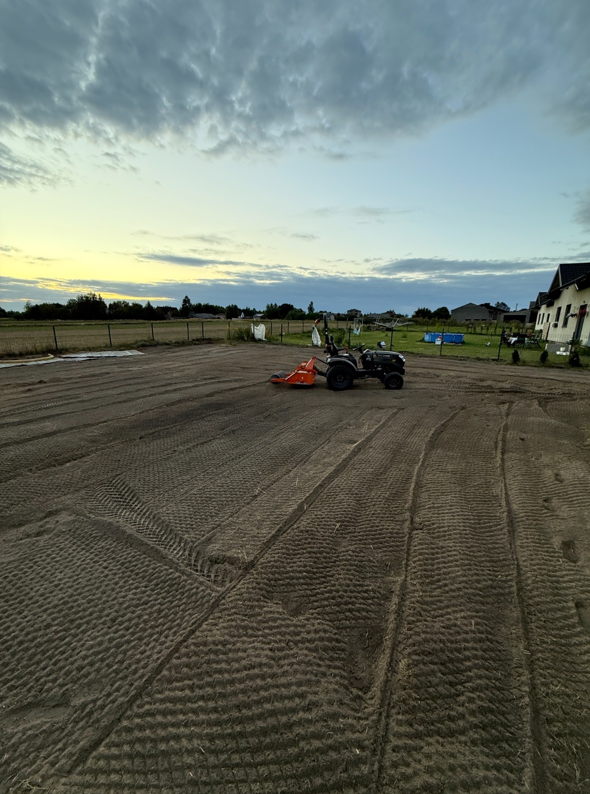 Niwelacja terenu pod trawnik: mały traktor z glebogryzarką spulchnia ziemię na działce. Widoczne ślady opon i spulchniona gleba. W tle dom i zachmurzone niebo.