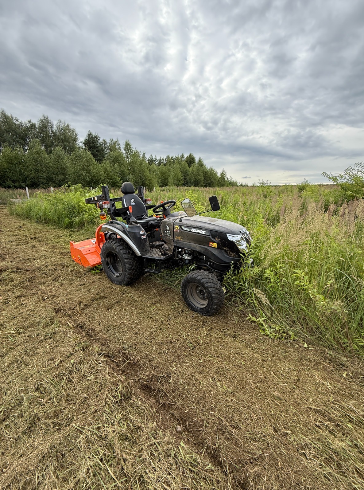 Czarny traktorek Solis z pomarańczowym kosiarką rotacyjną na polu z częściowo skoszoną trawą, w tle drzewa i pochmurne niebo. Widok z boku, lekko z góry.