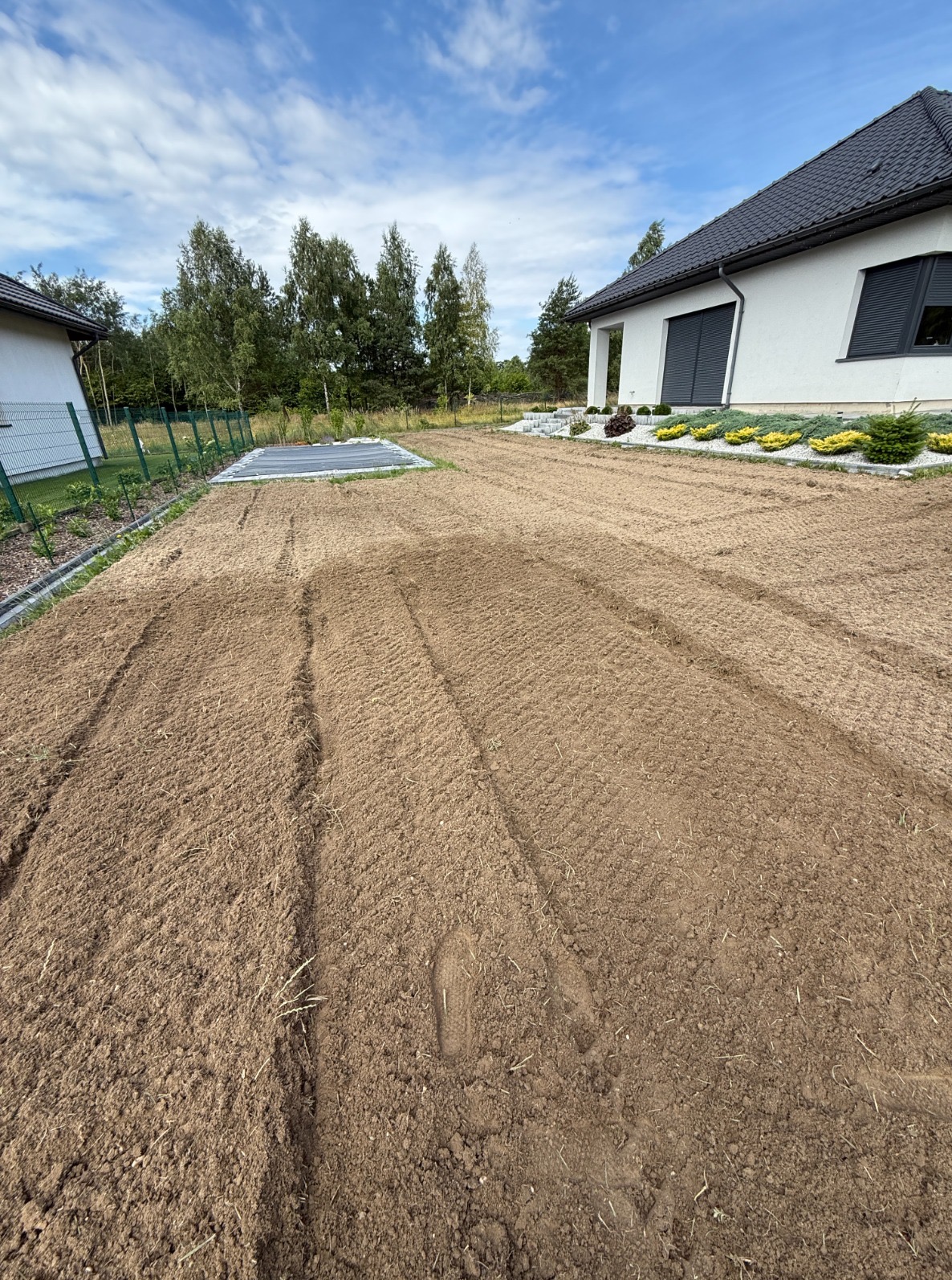 Przygotowany teren pod trawnik w Częstochowie. W tle dom z ciemną dachówką, widoczne ślady opon po maszynach. Grządki wyrównane, gotowe do siewu trawy.