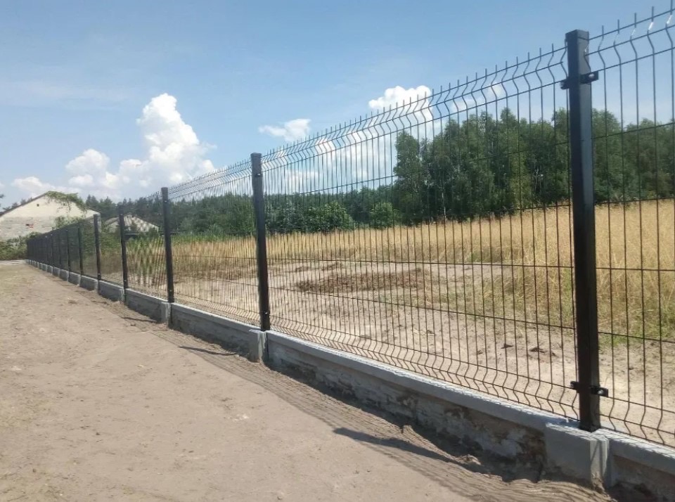 Długi, panelowy płot z siatki z podmurówką betonową, biegnący wzdłuż polnej drogi, z widokiem na zarośnięte pole i drzewa w tle, w słoneczny dzień.