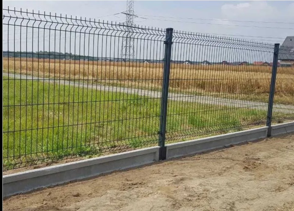 Nowoczesne ogrodzenie panelowe z podmurówką betonową na granicy działki z widokiem na łąkę i słup energetyczny w tle. Estetyczne i funkcjonalne rozwiązanie.