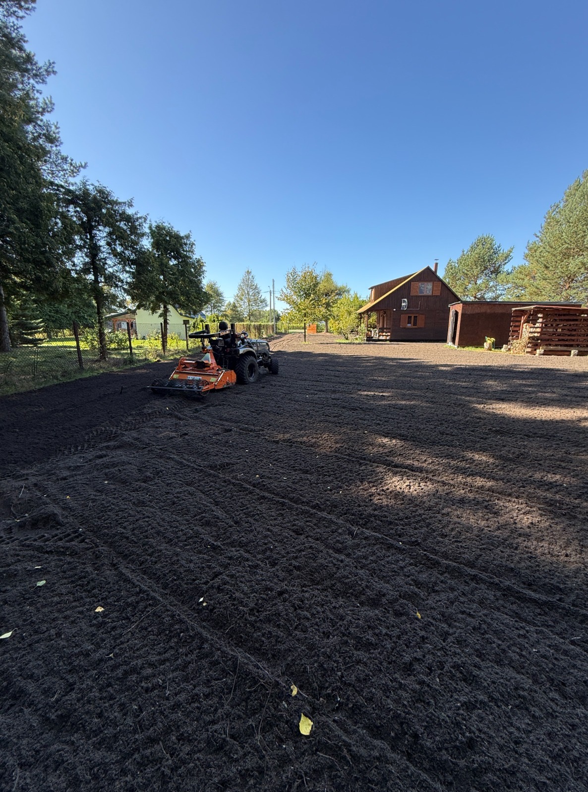 Niwelacja terenu pod trawnik: glebogryzarka wyrównuje ziemię na działce z domem i drzewami w tle, przygotowując podłoże pod założenie ogrodu.